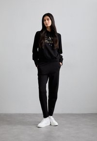 Čierna mikina s zlatým nápisom, skombinovaná s čiernymi nohavicami a bielymi teniskami. Materiál vyzerá mäkko; design je casual a priliehavý.