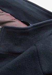 Giacca blu scuro con un esterno in lana testurizzata e una fodera interna liscia con un motivo bordeaux. Dettagli visibili della zip e del colletto.