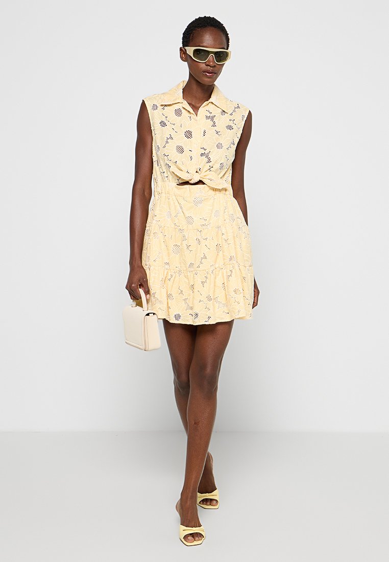 Vestido amarillo de volantes con un frente anudado, mangas cortas y patrón de encaje floral. Combinado con gafas de sol verdes y un bolso beige.