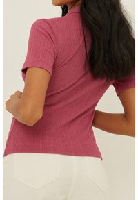 Krótki rękaw, żebrowana bluzka w kolorze magenta o dopasowanym kroju. Posiada kołnierzyk i lekko teksturowany materiał. Noszona z jasnymi spodniami.