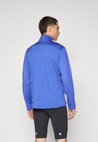 Blå sportjakke med høy krage, lange ermer og glatt tekstur, kombinert med svarte tettsittende shorts. Detaljer og sømmer er minimal.