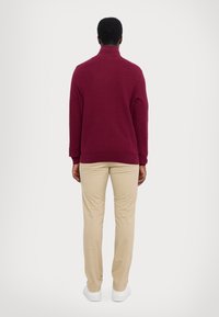 Bordeauxfarbener Pullover mit hohem Kragen, strukturierter Stoff. Beigefarbene Hose mit geradem Schnitt. Weiße Sneakers runden das Outfit ab.