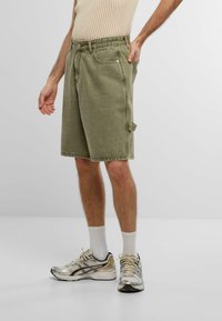 Shorts in denim verde oliva con taglio dritto, dotati di chiusura con bottone frontale, tasche laterali e un piccolo dettaglio a cerchio sull'orlo.