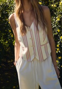 Gilet in lino a righe color crema con accenti rosa, design a scollo a V, cinque bottoni e tasche frontali; abbinato a pantaloni bianchi dalla vestibilità ampia.