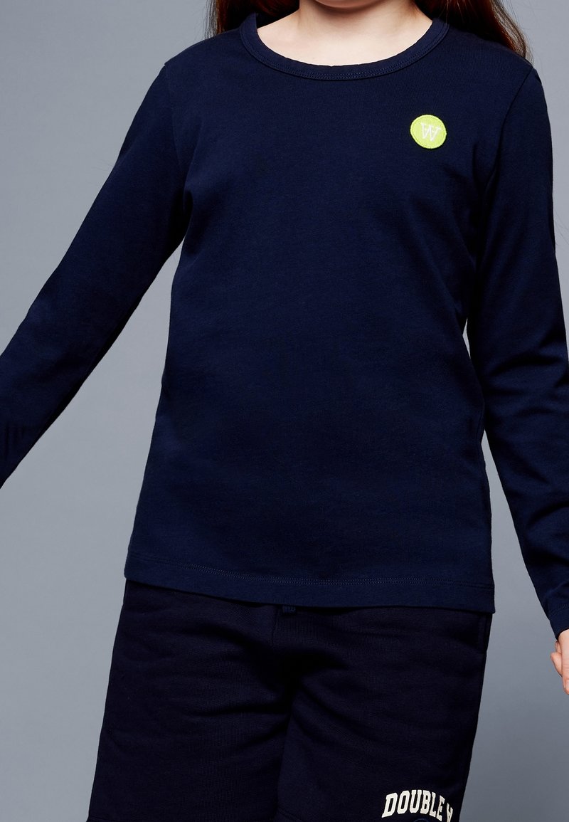 Dziecko w granatowej koszulce z długim rękawem z zielonym, okrągłym logo i czarnych szortach z białym napisem "Double X" na szarym tle.