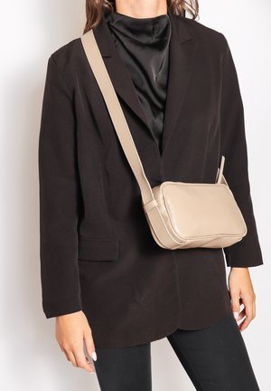 Sac en cuir marron clair en bandoulière de forme rectangulaire, texture lisse et bandoulière ajustable, porté sur un blazer noir et un haut.