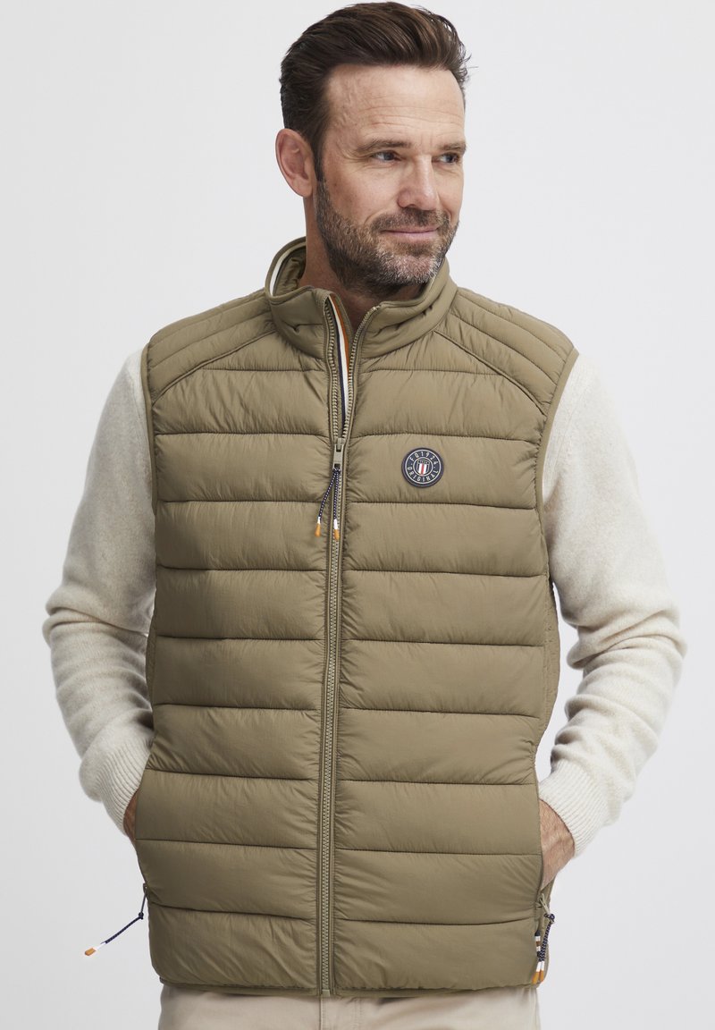 Olivgrüne gesteppte Weste mit hohem Kragen, Reißverschluss und Logopatch. Über einem cremefarbenen Langarmshirt getragen. Verfügt über seitliche Taschen.