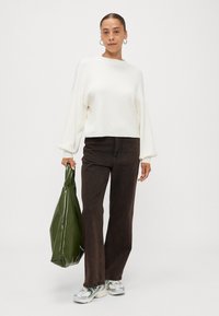 Weißer, kurz geschnittener Sweatshirt mit weiten Ärmeln, kombiniert mit dunkelbraunen, hochgeschnittenen Jeans. Auffällige grüne, glänzende Tragetasche und Sneakers.