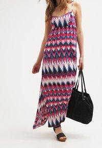 Robe longue à motif zigzag blanc, rose et bleu, avec des bretelles fines et une taille élastique. Associée à un sac fourre-tout noir et des sandales noires.