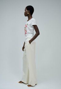 Camiseta blanca de algodón con estampado floral; pantalones crema de pierna ancha; pendientes de aro dorados; modelo posando de lado contra un fondo neutro.