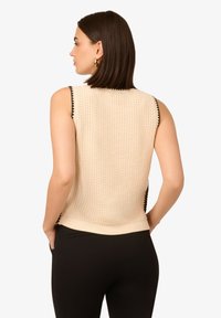 Beige ärmelloses Top mit einem strukturierten Strickmuster, das schwarze Wellenkanten entlang der Armausschnitte und Seitennähte aufweist.