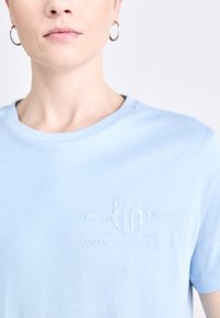Gaiši zils kokvilnas T-krekls ar apaļu kakla izgriezumu un teksturētu izšūtu logo krūtīs, kas ietver tekstu "GANT AMERICAN SPORTSWEAR."