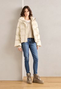 Μπεζ φουσκωτό μπουφάν με ψηλό γιακά, συνδυασμένο με ένα ανοιχτό πλεκτό πουλόβερ, μπλε τζιν και καφέ μπότες με κορδόνια πάνω σε ξύλινο πάτωμα.