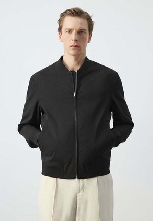 Man with light skin and short brown hair wearing a black zip-up jacket and light beige trousers, standing against a plain background.