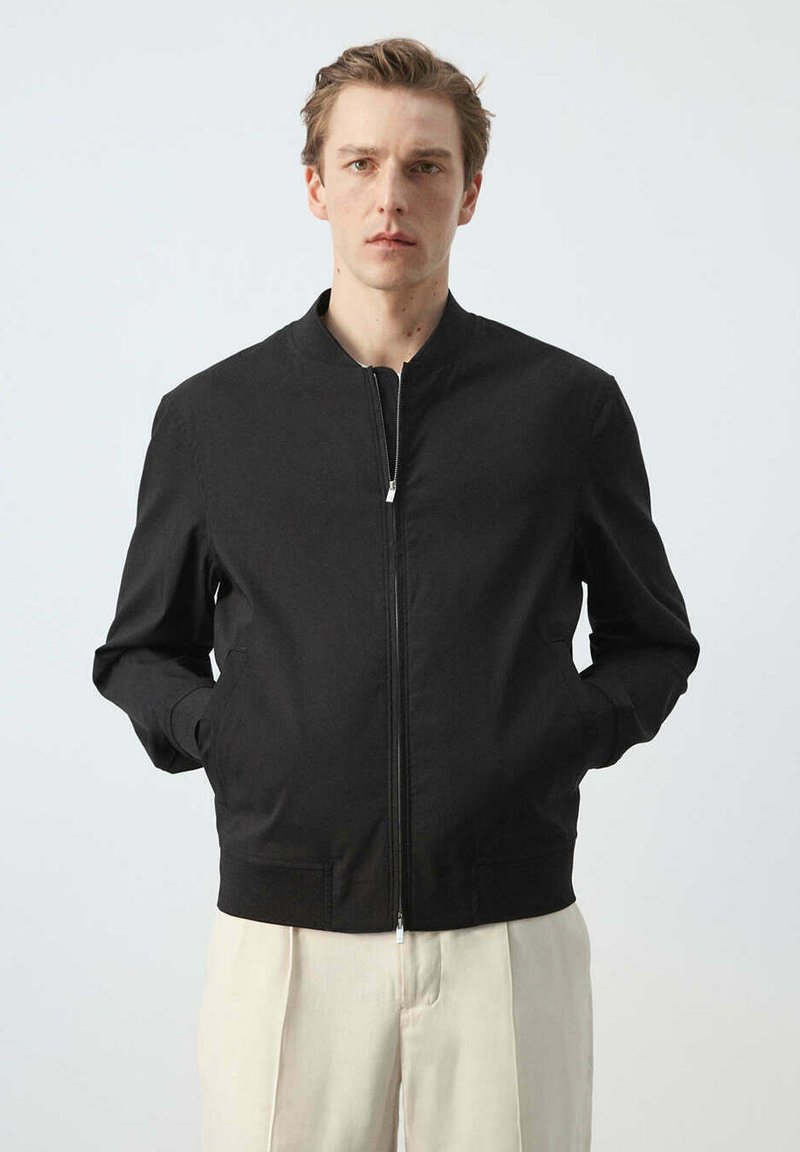 Homme à la peau claire et aux cheveux courts bruns, portant une veste noire zippée et un pantalon beige clair, debout devant un fond uni.