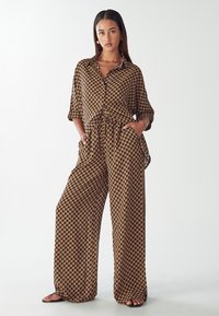 Brown patterned jumpsuit with a loose fit, short sleeves, a collar, elastic waist, and side pockets. Features a geometric design.