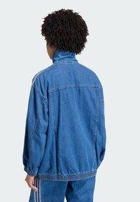 Veste en denim bleu avec un col montant, des poignets élastiqués et une taille cintrée. Présente des coutures visibles et une coupe décontractée.