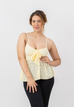 Frau mit lockigem Haar, die ein blassgelbes Spitzen-ärmelloses Top und schwarze Hosen trägt, posiert mit einer Hand in der Taille und der anderen entspannt.