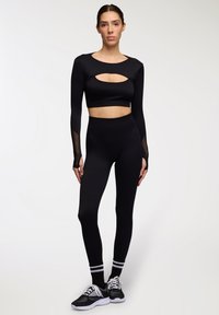 Completo da allenamento nero composto da un crop top a maniche lunghe con aperture e dettagli in rete, abbinato a leggings a vita alta e scarpe sportive.