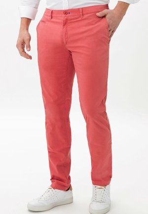 Mand iført koralfarvede, slanke bukser, hvide sneakers og en hvid skjorte med knapper, med den ene hånd i lommen.