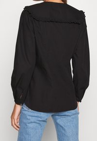 Blouse noire avec un col volanté, de longues manches bouffantes et des poignets boutonnés. Le tissu est lisse avec une coupe cintrée, portée avec un jean bleu clair.