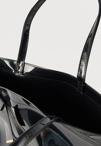 Γυαλιστερή μαύρη τσάντα ώμου από λουστρίνι με δύο χερούλια, που δείχνει λεπτομερή ραφή και ανοιχτό επάνω μέρος σε απλό φόντο.