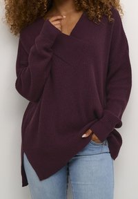 Mysig, oversized, ribbad stickad tröja i djup lila med V-hals och sidoflikar, ihop med ljusblå jeans.