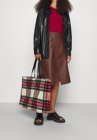 Musta nahkapaitatakki, punainen paita, ruskea nahkainen kietaisuhame ja mustat sandaalit. Kantaa ruudullista kangaskassia, jossa on punainen, musta ja kerma.