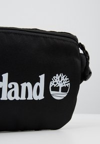 Sac en tissu noir avec logo Timberland blanc, comprenant texte et symbole d’arbre, visible sur un fond clair uni.