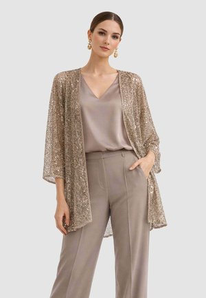 Donna con blusa di raso beige, pantaloni a vita alta abbinati e cardigan aperto in paillettes dorate, con orecchini vistosi.