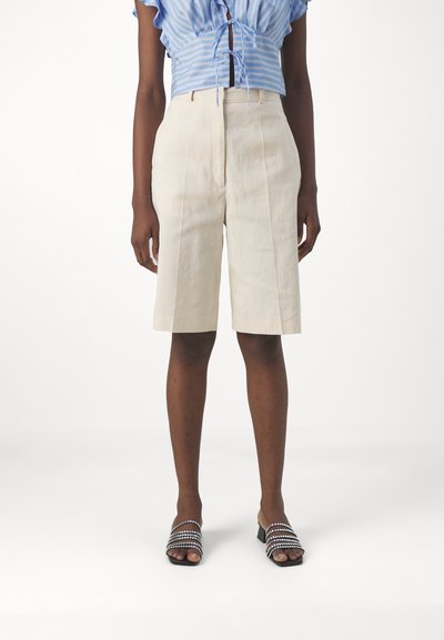 Bermudas en lin beige avec des plis sur le devant, associées à un chemisier rayé bleu et blanc. Portées avec des sandales noires ornées de strass.