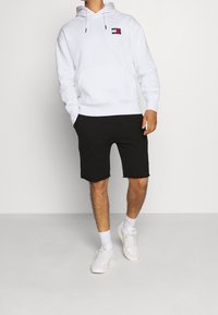 Person wearing a white Tommy Jeans hoodie, black shorts, white socks, and white sneakers standing against a plain background.