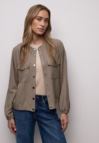 Beige leichte Jacke mit Druckknöpfen, zwei Brusttaschen, elastischen Bündchen und einem leicht verkürzten Saum, kombiniert mit einem beigen Oberteil und blauen Jeans.