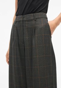 Sive karirane hlače od teksturirane tkanine s narančastim naglascima, koje imaju plisirane detalje i ravni kroj. Vidljiva ruka u džepu.