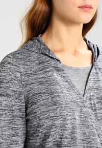 Žena v šedé melírované pletené mikině s límcem a světle šedou košili pod ní, zobrazená od krku až po hruď.