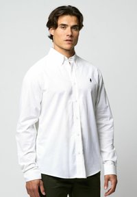 Man in een geknoopte witte blouse met lange mouwen en kraag, met donkere broek, staande tegen een effen lichtgrijze achtergrond.