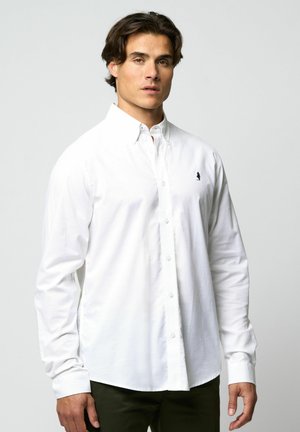 Hombre vestido con una camisa blanca de mangas largas abotonada con cuello y pantalones oscuros, de pie contra un fondo gris claro liso.