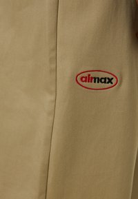 Beige fabric with a smooth texture. Features an embroidered red and black logo. Tailored fit with clean lines and no patterns or accents.