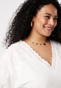 Weiße Bluse mit bestickten Akzenten und einem gewellten V-Ausschnitt. Halskette mit goldfarbenem Herzanhänger und großen goldenen Creolen. Dunkel lockiges Haar.