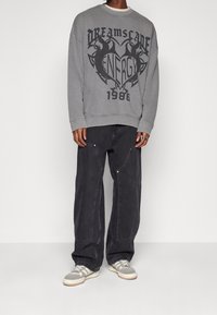 Sudadera gris de gran tamaño con estampado gráfico negro, combinada con jeans amplios de color gris oscuro que tienen bolsillos laterales y detalles en plata. Las zapatillas son grises con rayas.