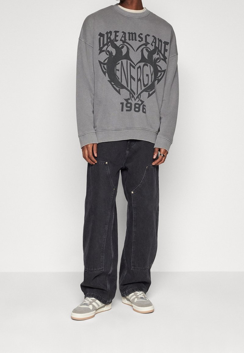 Sudadera gris de gran tamaño con estampado gráfico negro, combinada con jeans amplios de color gris oscuro que tienen bolsillos laterales y detalles en plata. Las zapatillas son grises con rayas.