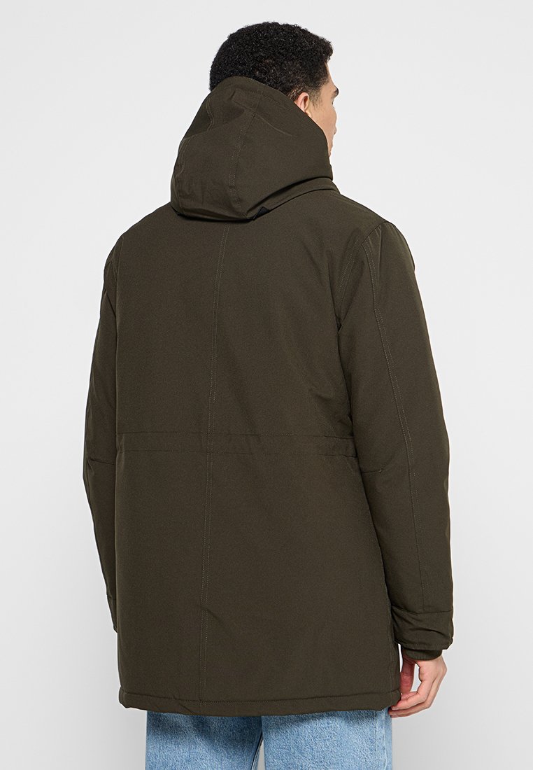 Veste matelassée vert foncé avec capuche, présentant une coupe droite et des détails de couture visibles. Texture lisse et design fonctionnel.