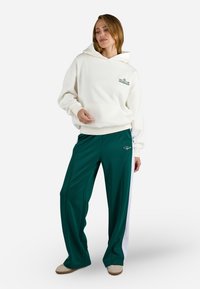 Felpa bianca con cappuccio e tasca frontale; abbinata a pantaloni verdi con righe bianche laterali e un logo discreto. Tessuto morbido.