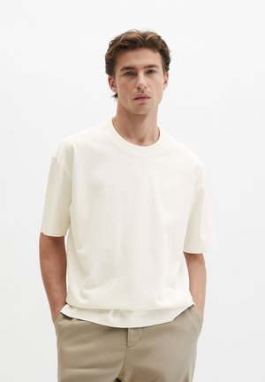 Junger Mann mit hellbraunem Haar, trägt ein schlichtes weißes Kurzarmhemd und beige Hosen, steht mit den Händen in den Taschen vor einem weißen Hintergrund.