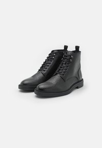 Botas de cuero negras hasta el tobillo con una superficie texturizada, punta redondeada, diseño con cordones y una suela de goma resistente. Incluye una pestaña para facilitar el uso.