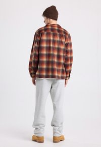 Camicetta a quadri in tonalità di rosso, arancione e marrone, abbinata a pantaloni della tuta grigi. L'outfit include un berretto lavorato a maglia marrone e stivali color sabbia.