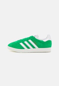 Green suede sneaker with white three-stripe design, white heel, and textured rubber sole. Features gold "Gazelle" branding on the side.