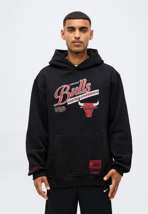 Jeune homme portant un sweat à capuche noir des Chicago Bulls avec des logos rouges et blancs et du texte, debout devant un fond clair uni.