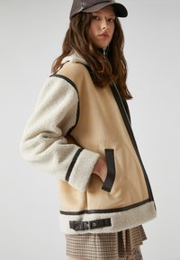 Beige- und cremfarbene Jacke aus Kunstfell und Wildleder, mit schwarzen Akzenten, Reißverschluss vorne und zwei Seitentaschen.