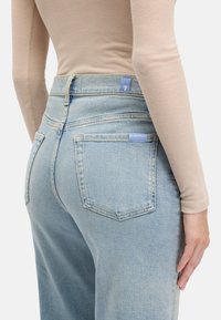 Jean bleu clair taille haute avec une coupe droite, comprenant cinq poches, une étiquette bleue à l'arrière, et une légère usure subtile.